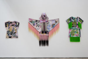 3 textile pieces that look like shirts or capes are displayed side-by-side. They are all ornately detailed with patches and hair.